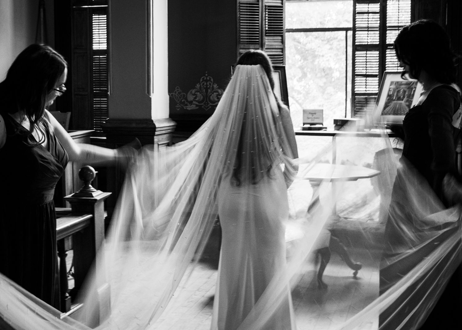 Black and white photo of wedding preparations with flowing veil in natural window light.
