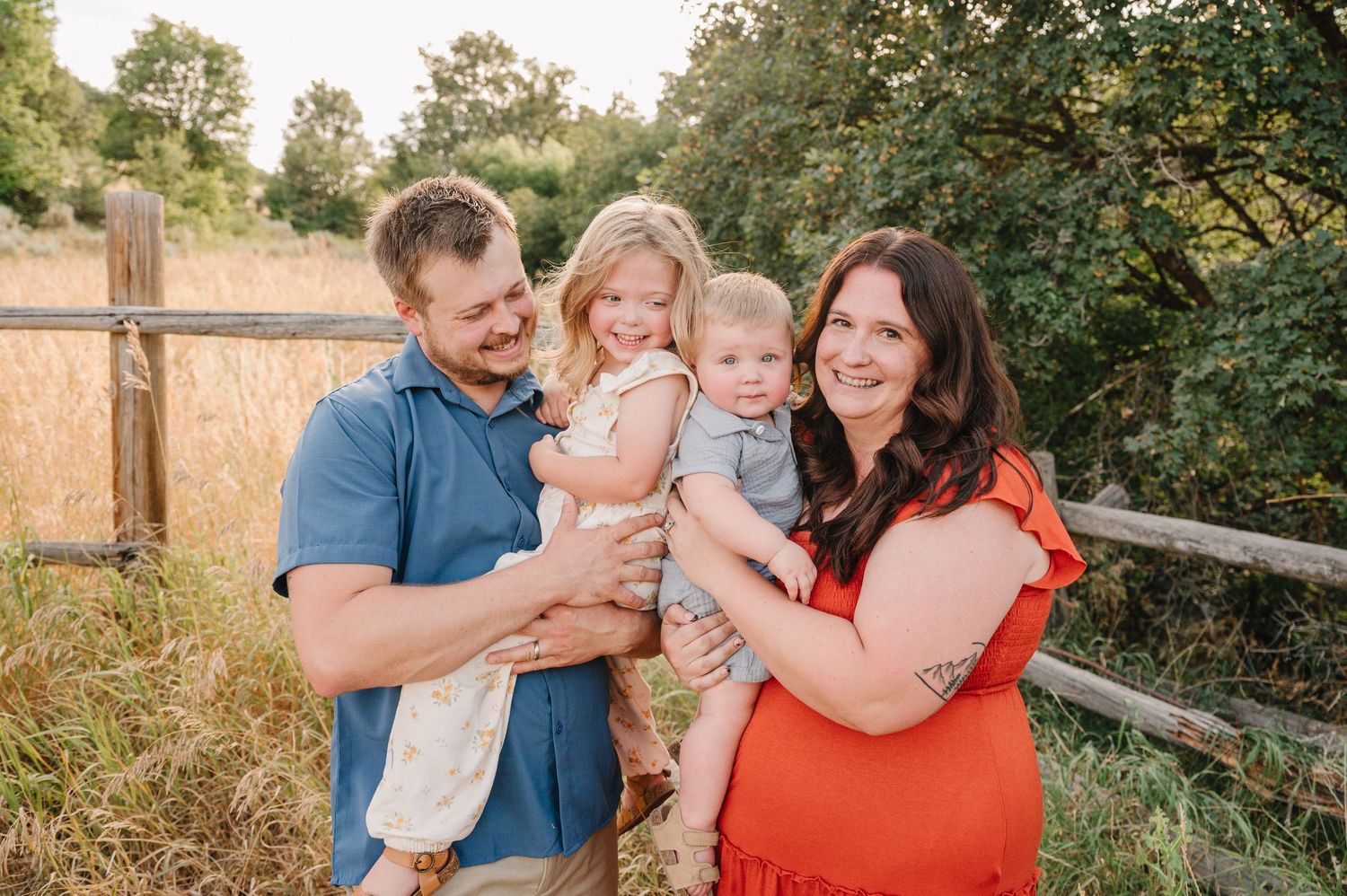 A family wearing blue and coral outfits embraces while posing for outdoor photos in a rustic setting at sunset.