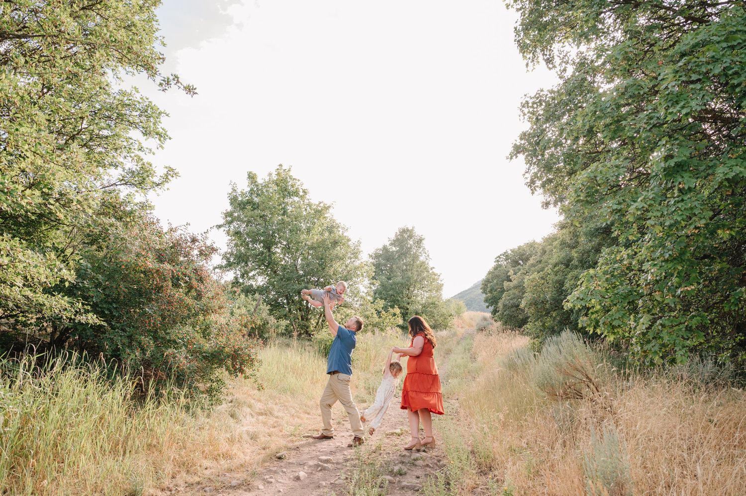 Two people walk hand in hand down a dirt path surrounded by tall grass and trees on a sunny summer day.