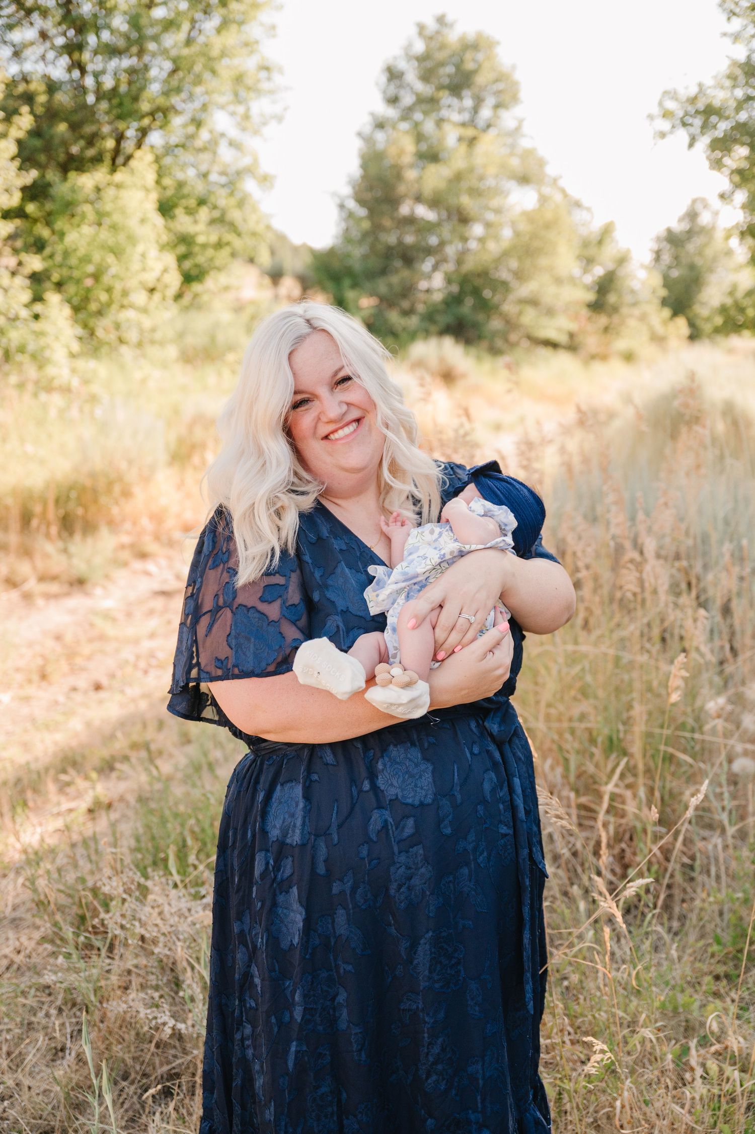 A mother in a navy blue dress holds and embraces a newborn baby in a sunlit outdoor field setting.