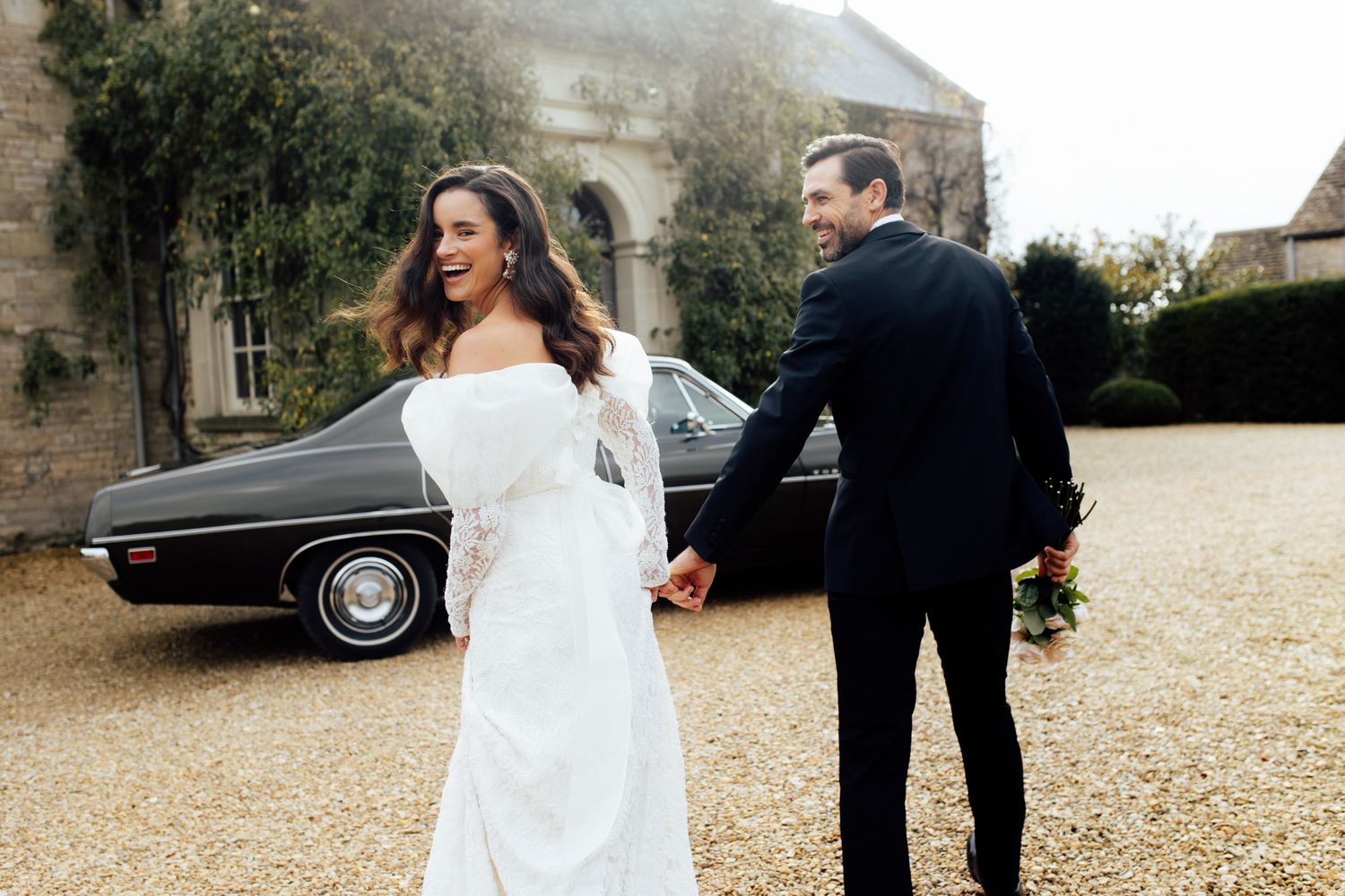 Newlywed couple walking together on a gravel path with vintage car in background. Euridge Manor Wedding.