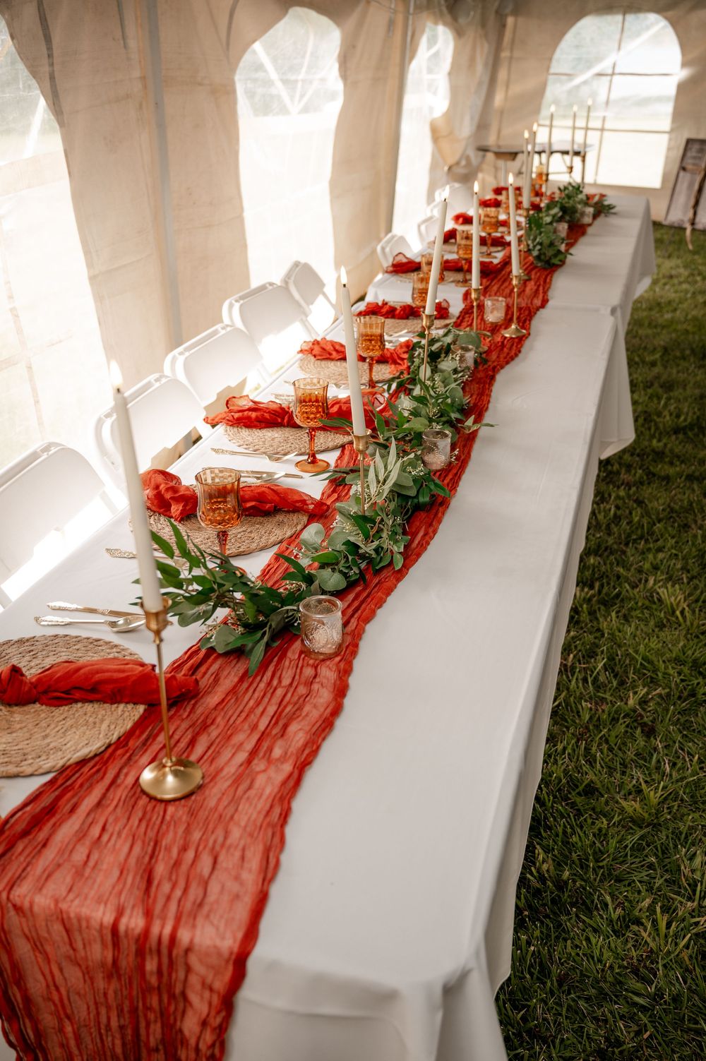 Wedding reception head table with red gauze runner, gold candles, and greenery under tent.