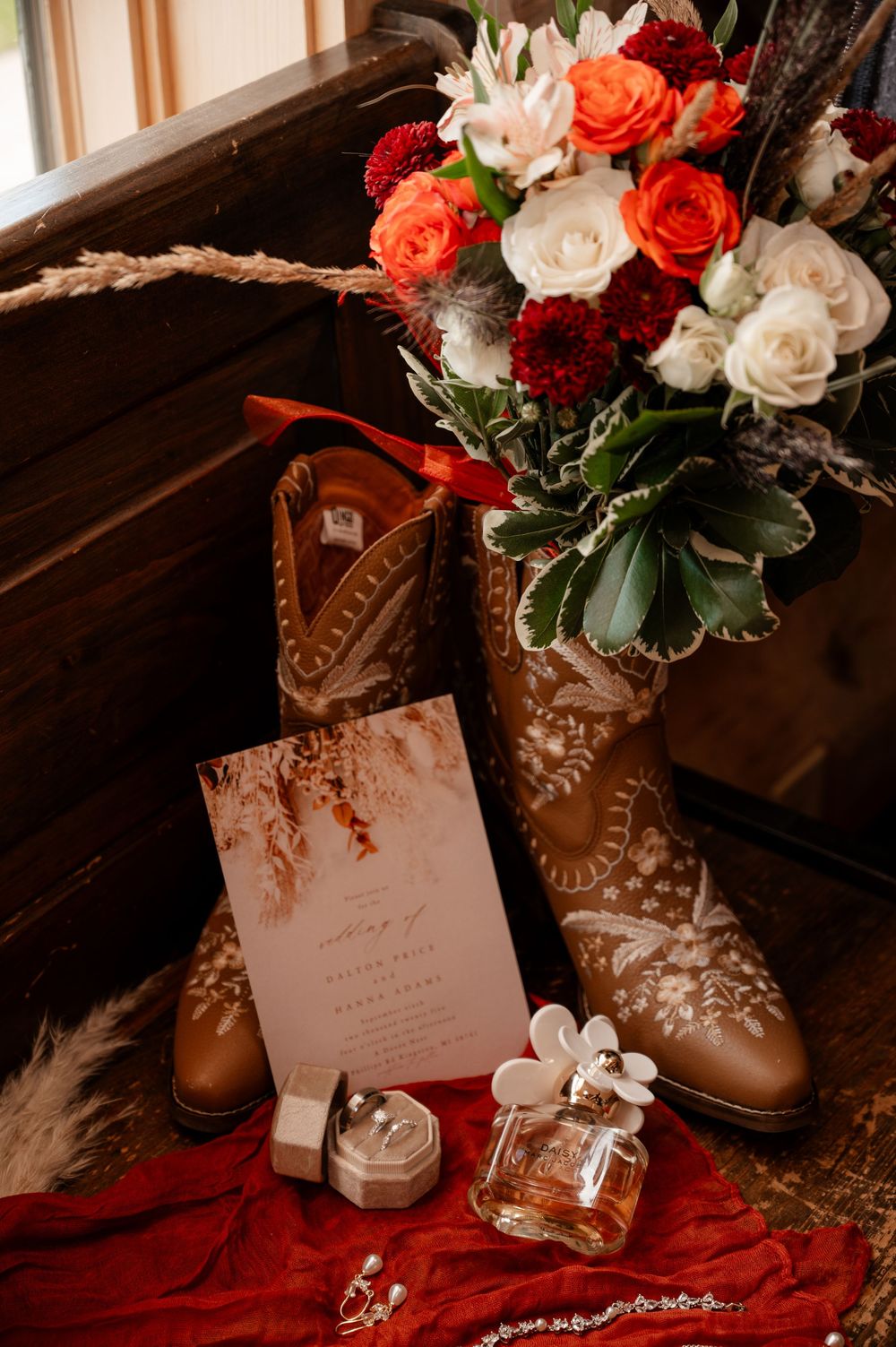 Cowboy boots holding bridal bouquet of red and white roses beside wedding invitation and perfume.