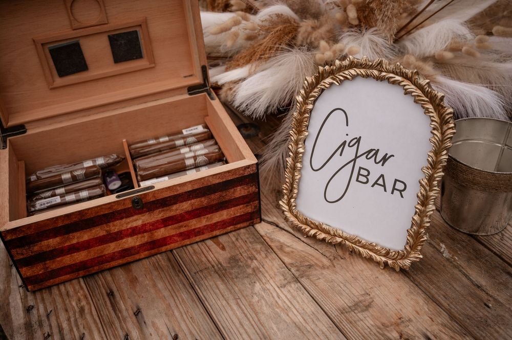 Wooden cigar humidor with cigars beside gold framed Cigar Bar sign on rustic wood floor.