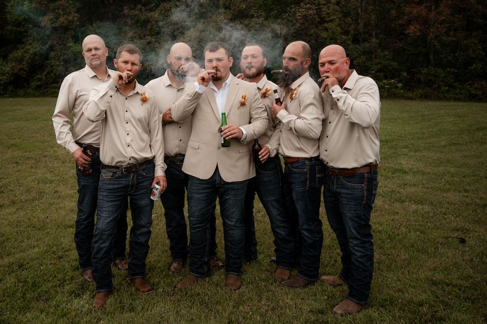 Groom and groomsmen in tan blazers and jeans smoking cigars outdoors at fall wedding.