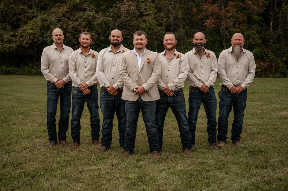 Groom with groomsmen wearing tan blazers and dark jeans posing in a green field.