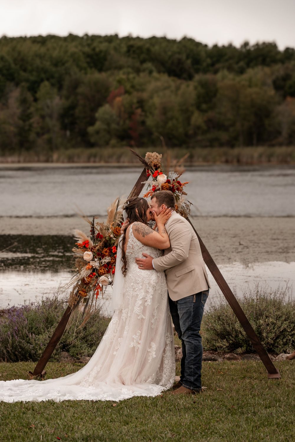 Bride and groom share first kiss at triangular wooden arch with fall floral decor by lake.