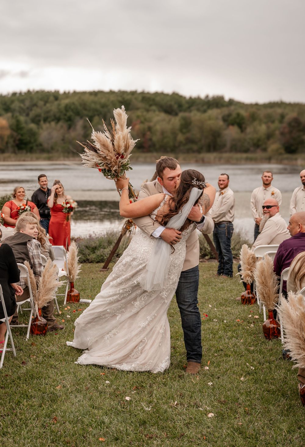Bride and groom share romantic dip kiss at outdoor lakeside wedding ceremony with pampas grass aisle decor.