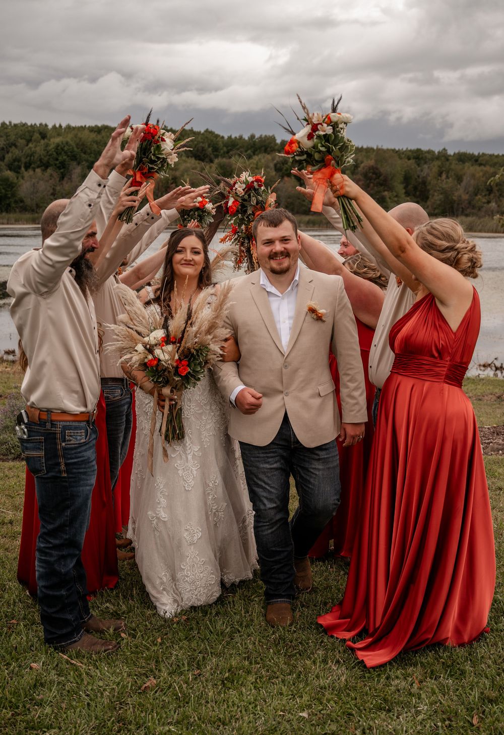 Wedding party in red dresses and tan suits raise bouquets over smiling bride and groom by lakeside.
