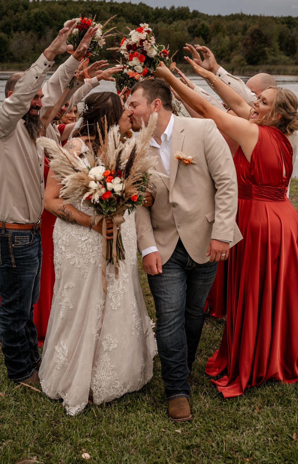Bride and groom kiss under arch of raised hands and bouquets surrounded by wedding party outdoors.