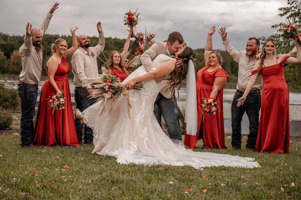 Groom dips bride for kiss as excited wedding party celebrates behind them at outdoor lakeside venue.
