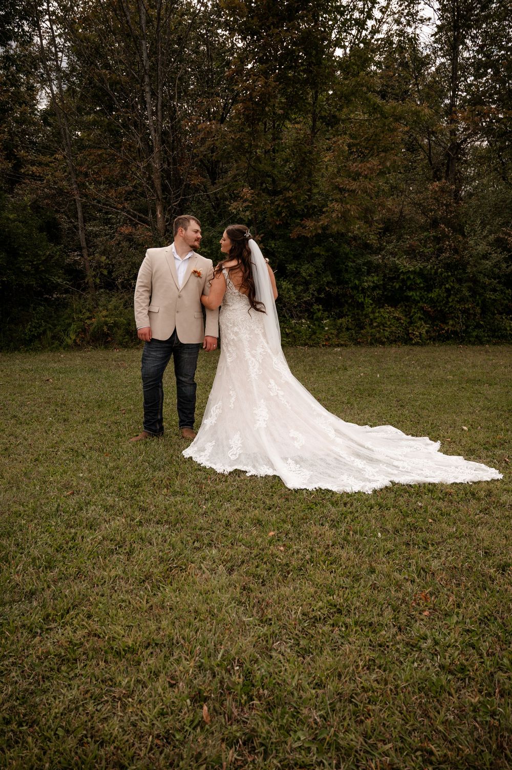 Bride in lace gown with cathedral train and groom in tan suit stand in green field near forest.