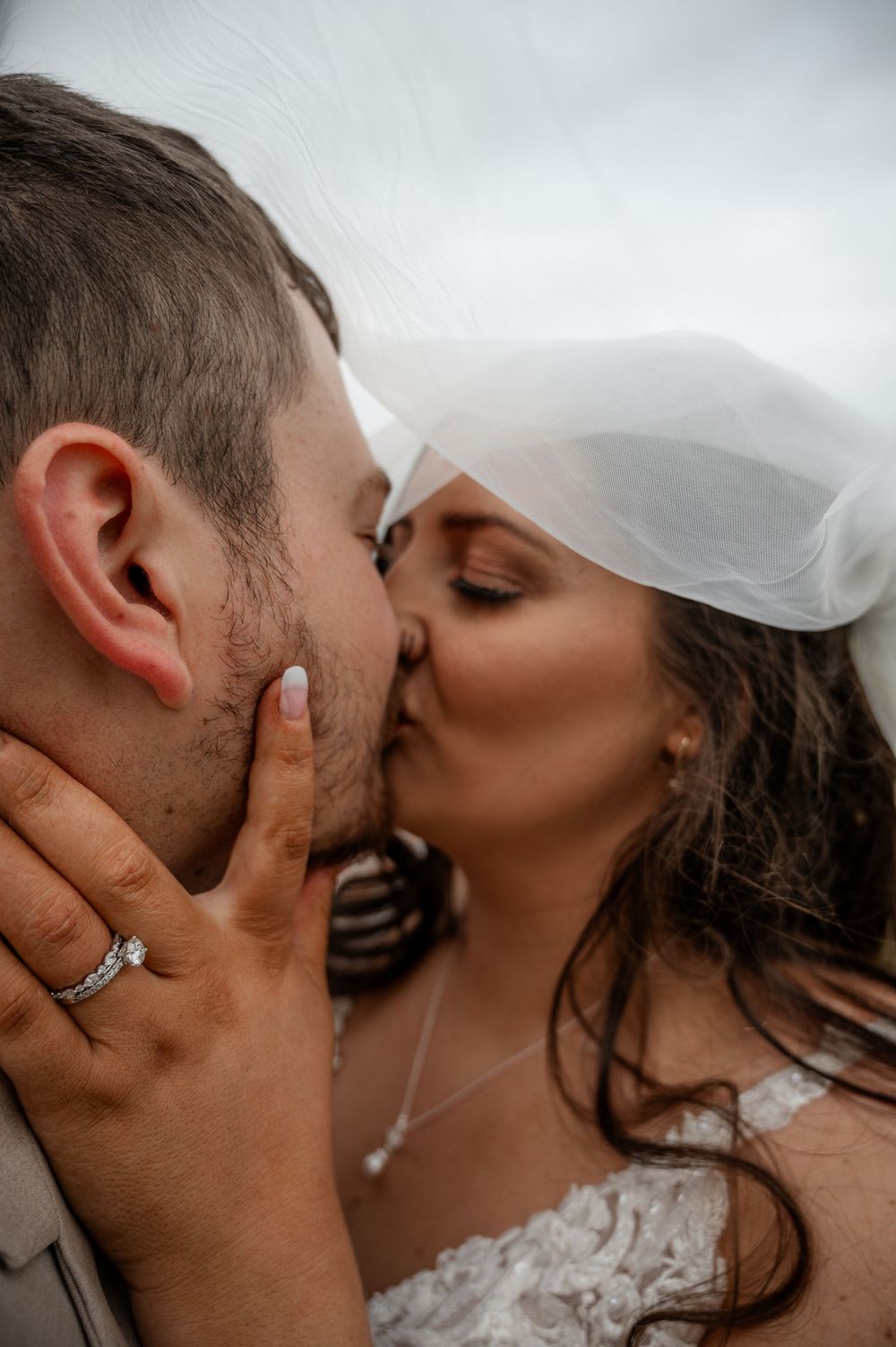 Close-up of bride and groom kissing under veil showing diamond engagement ring and wedding band.