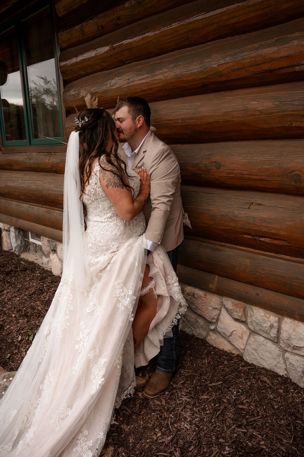 Groom in tan suit kisses bride in lace gown against rustic log cabin wall at wedding venue.