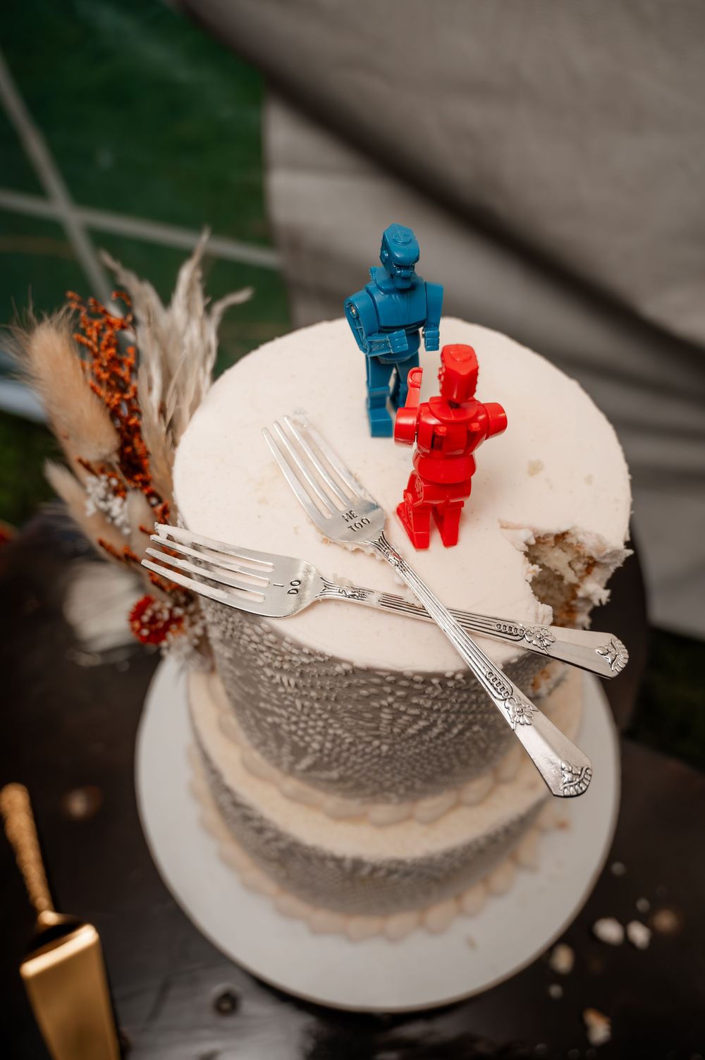 Two-tier white wedding cake topped with blue and red robot figurines and vintage silver forks.