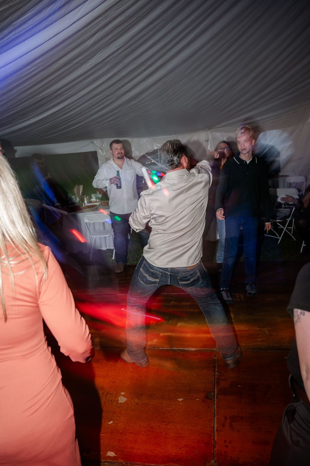 Wedding reception guests dancing energetically under tent with colorful lights during celebration.