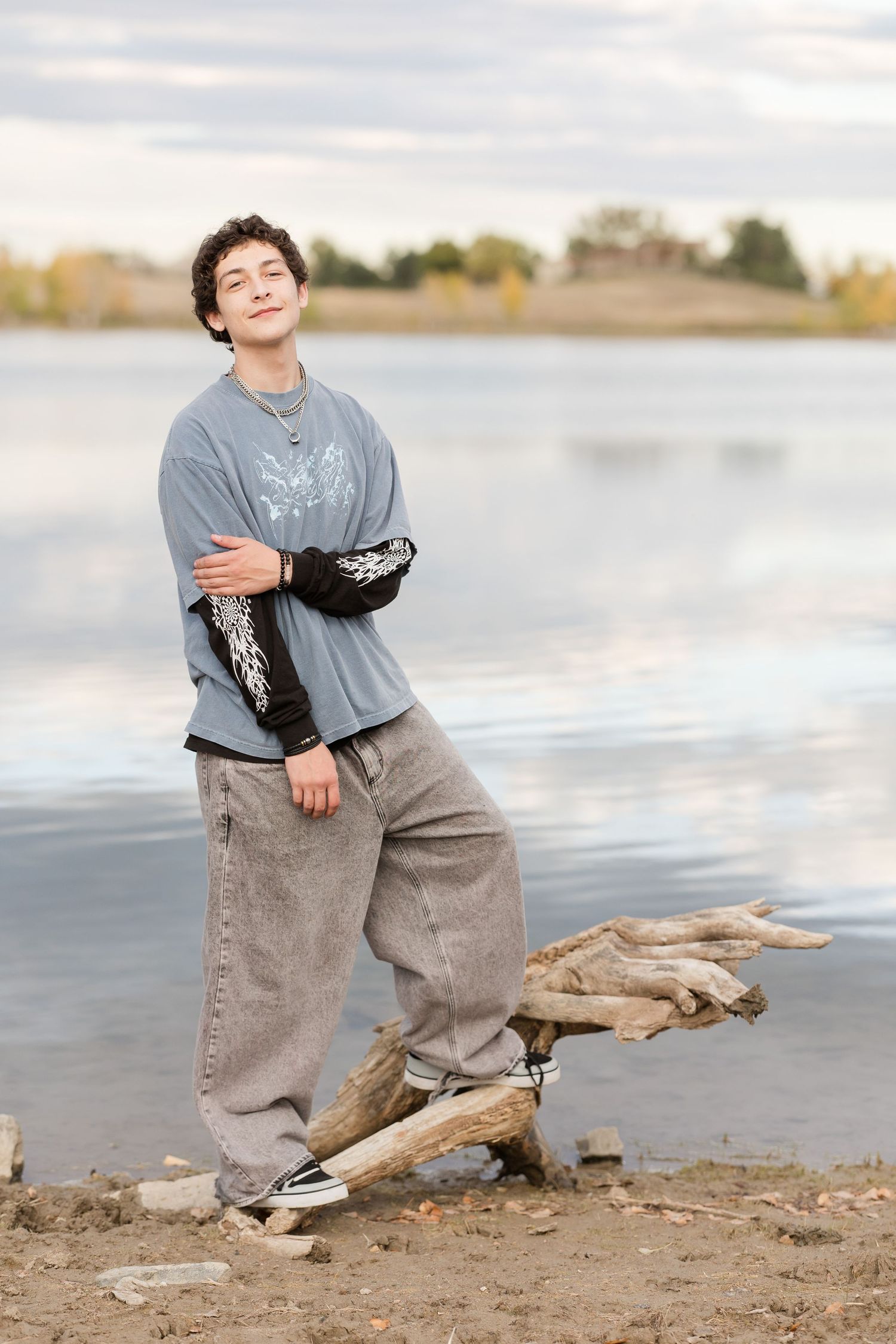 A figure in a gray t-shirt and jeans poses playfully near driftwood on a beach by a lake at sunset.