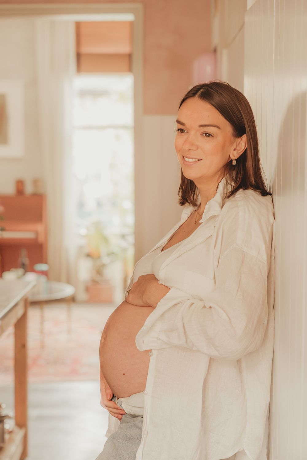 Expectant mother in white button-down shirt poses near window in warm natural light during maternity photoshoot.