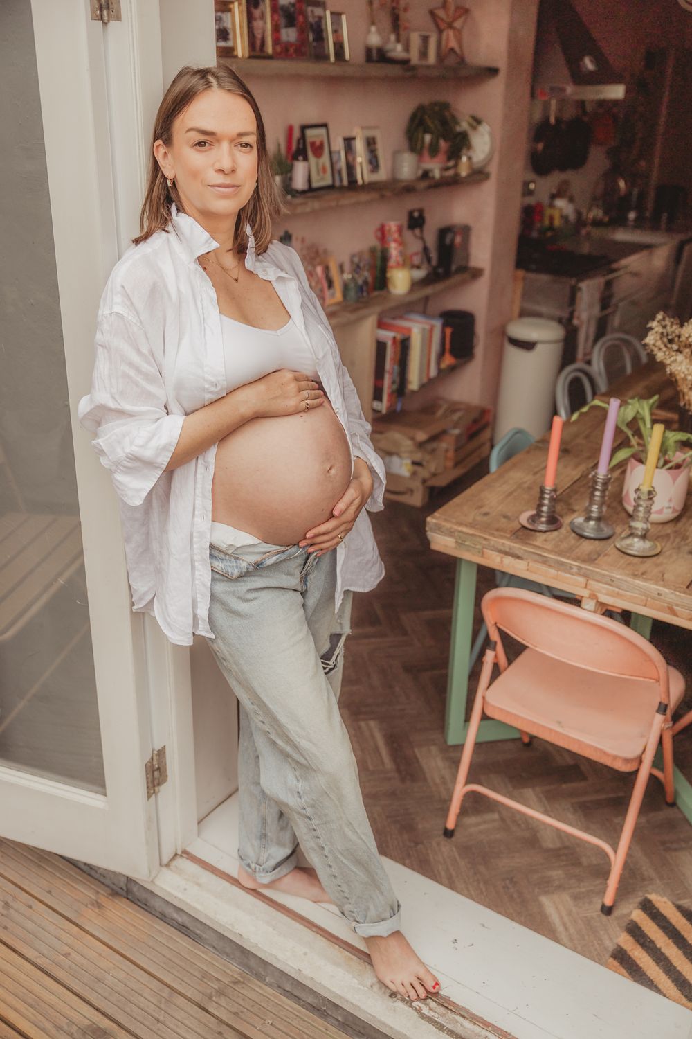 A pregnant woman in a white shirt and gray overalls sits on a yoga mat in a cozy home setting.