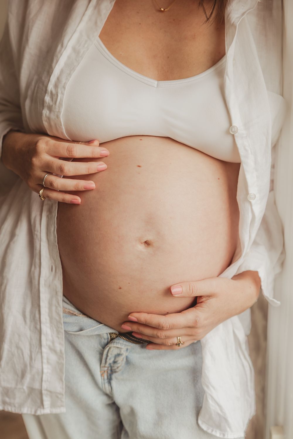 Close-up of pregnant belly being cradled by hands, wearing white top and light denim jeans.