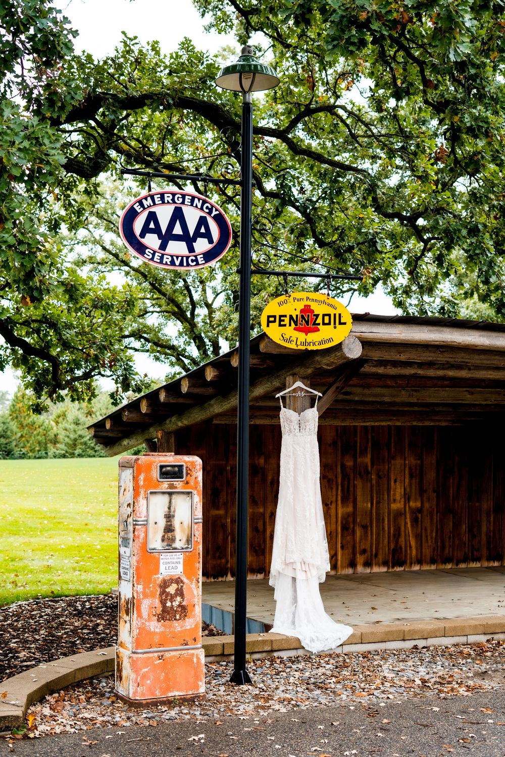 A wedding dress displayed outdoors near vintage AAA and Pennzoil signs against a wooden structure.