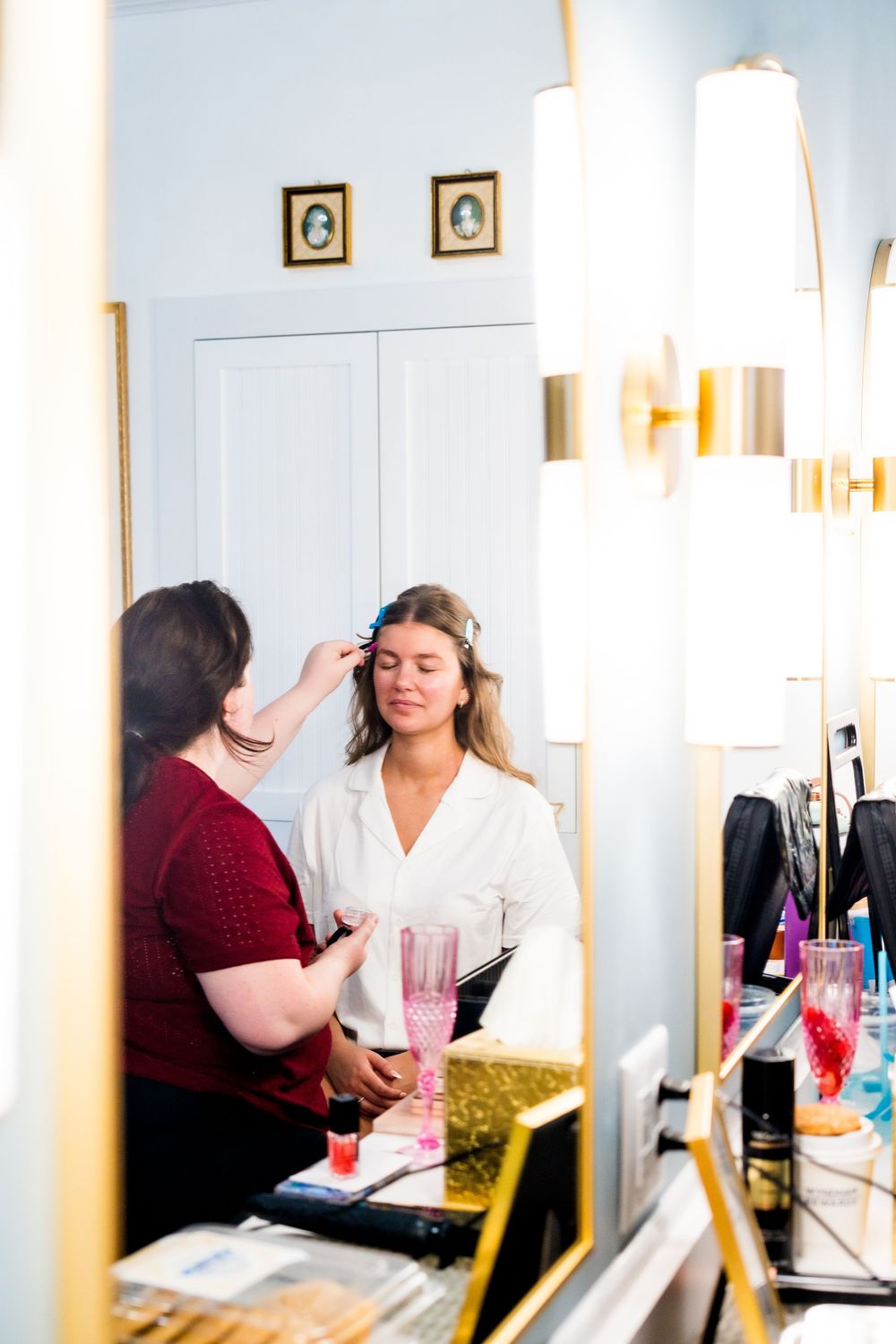 Bright sunlit room with makeup stations and mirrors showing wedding day preparations.