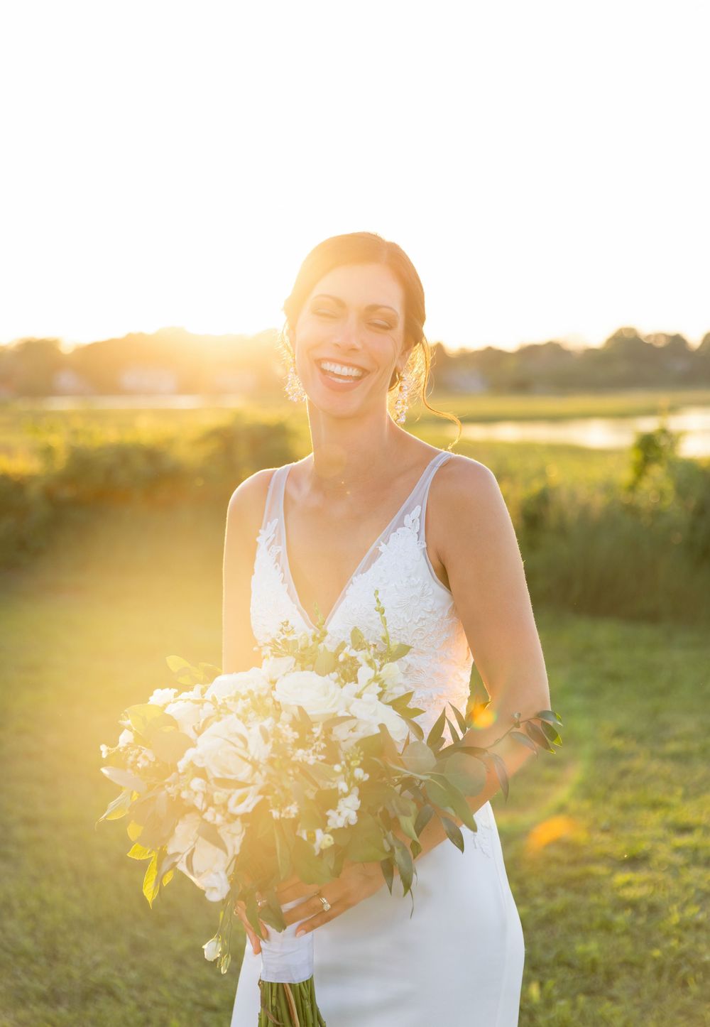 Bride holding white floral bouquet laughs joyfully in golden sunset light.
