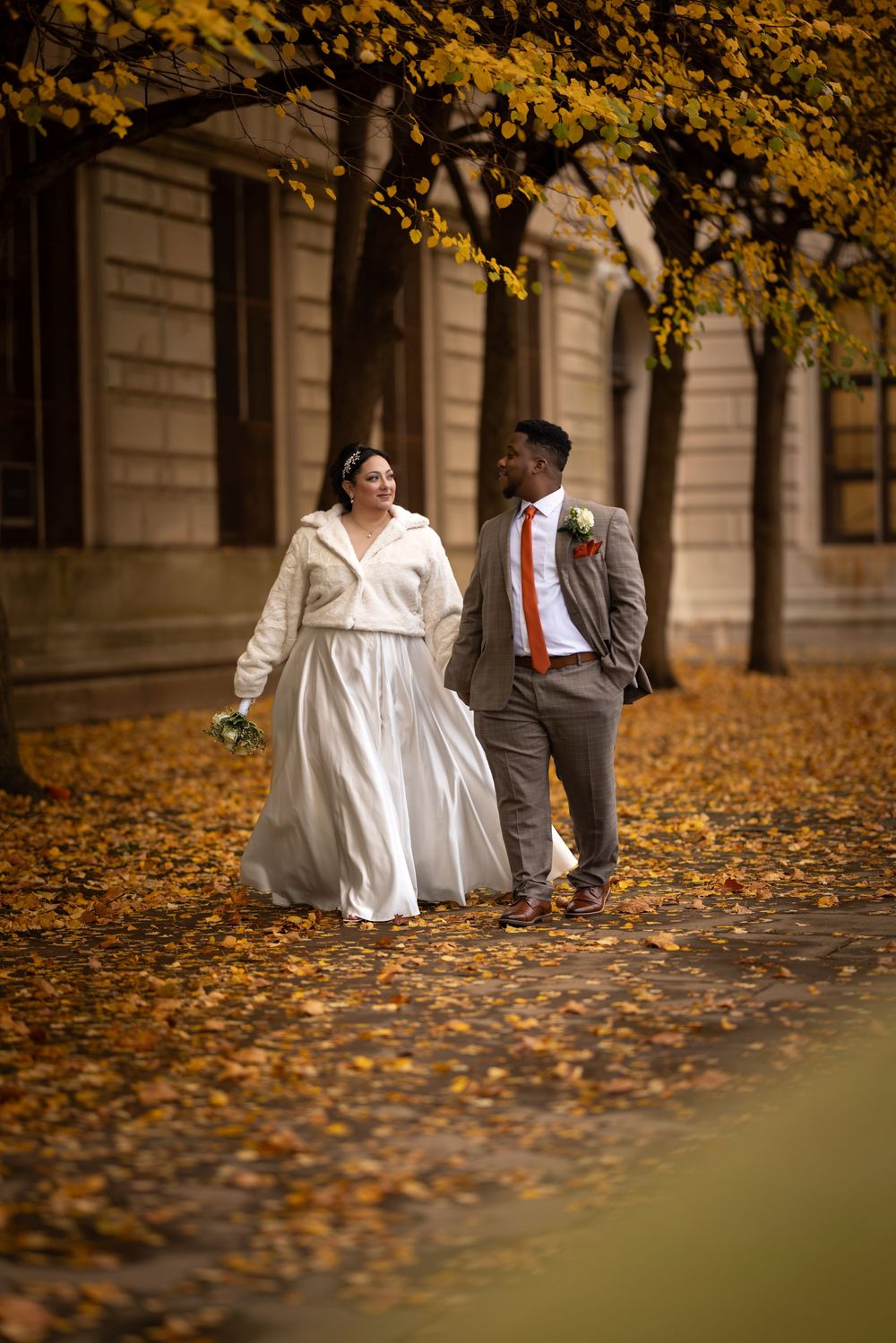 A couple walks together on a romantic autumn path covered in golden leaves while holding hands.