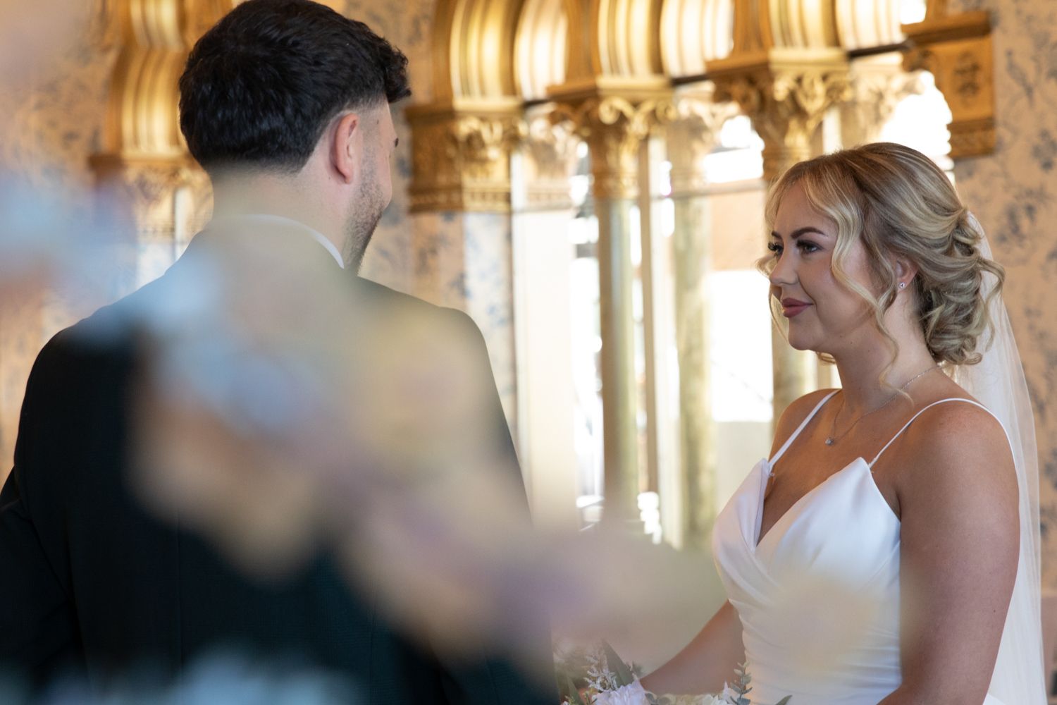 A couple exchanges vows during their wedding ceremony in an ornate venue with golden architectural details.