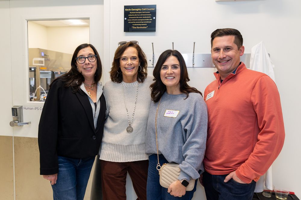 Four colleagues stand together smiling in an office setting wearing casual business attire.