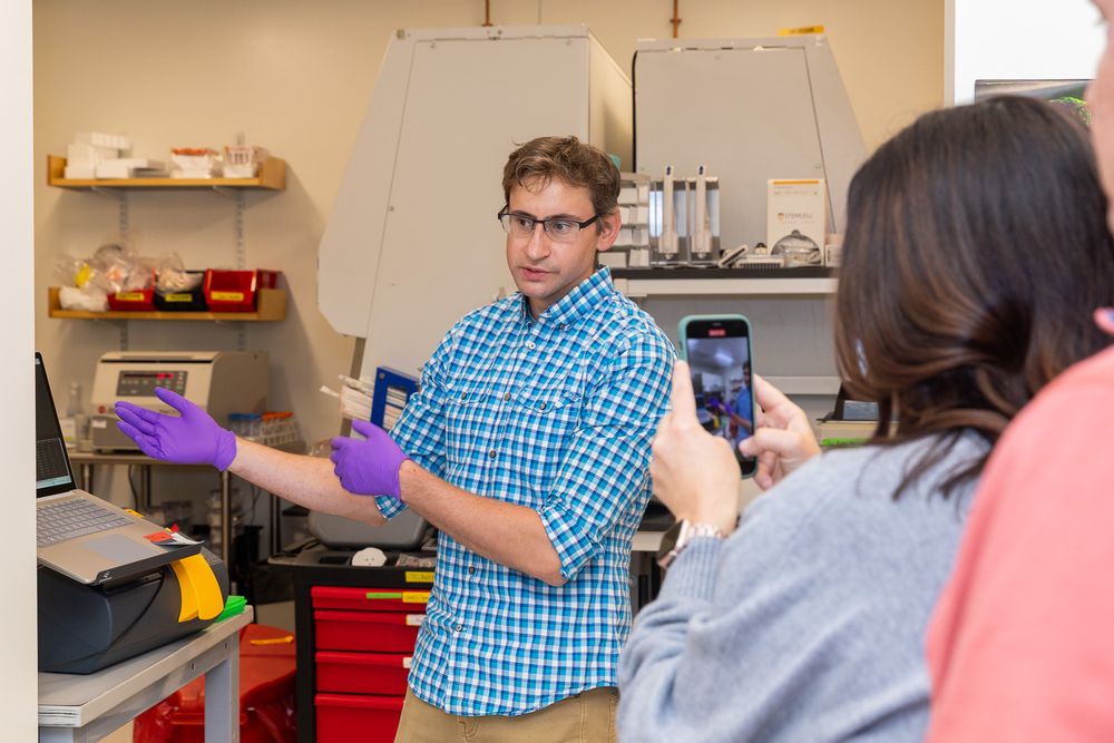 Laboratory researcher wearing purple gloves explains scientific procedure while someone takes a photo on smartphone.