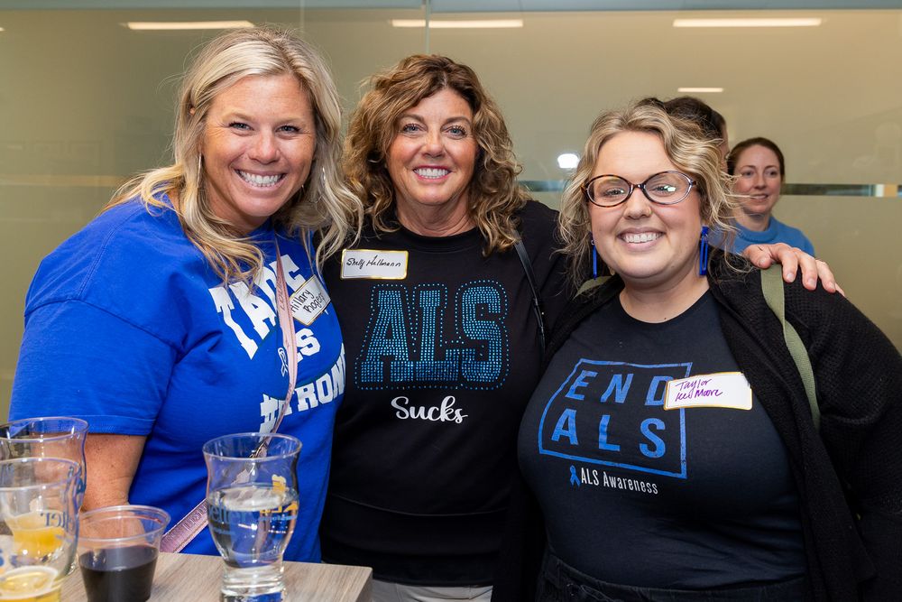 A group of people wearing blue and black t-shirts smile together at a casual gathering or event.