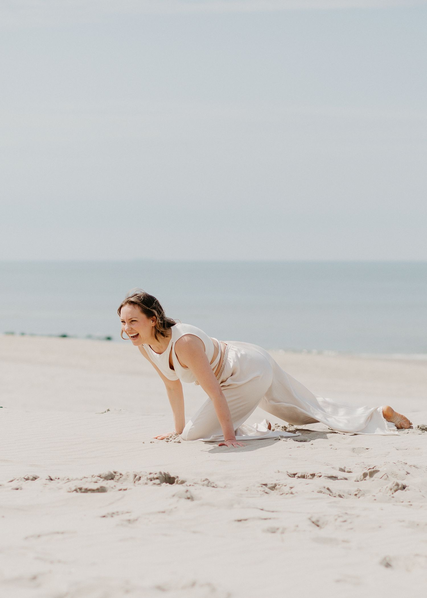 A joyful woman in a white dress crawling playfully on a sandy beach with the ocean in the background.