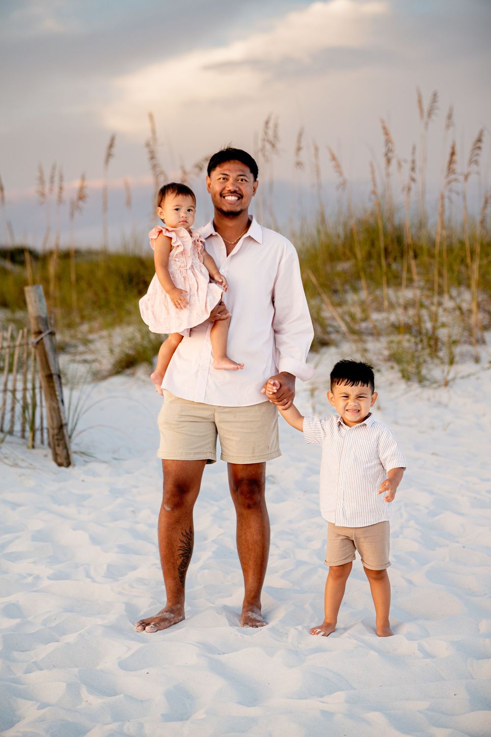 A family stands together on a white sandy beach with sea oats swaying in the background during sunset.