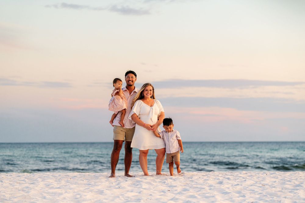 Family in white outfits walking together on white sandy beach during sunset.