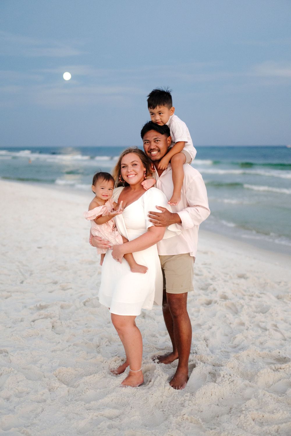 Family enjoying playful moments together on a white sandy beach with ocean waves in the background.