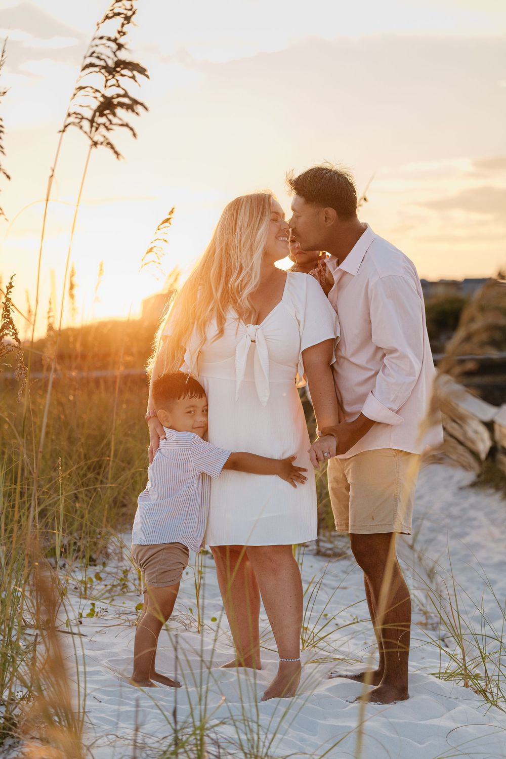 Romantic couple in white outfits sharing intimate moments on a sandy beach at sunset with sea oats in the foreground.