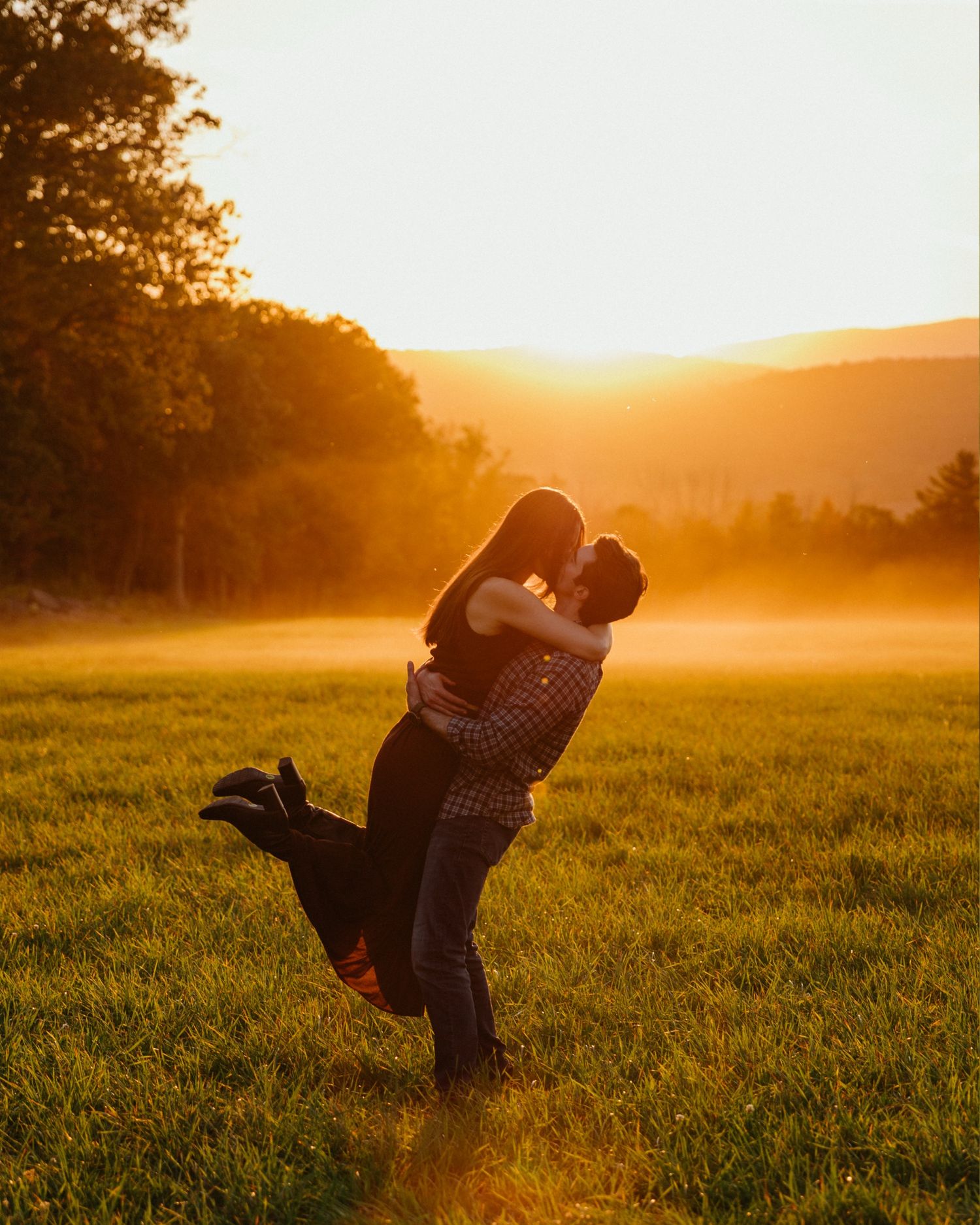 Silhouetted couple embracing in a misty meadow at golden sunset with sunlight streaming through trees.