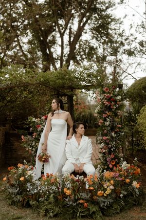 A romantic garden wedding scene with two people in white attire seated among blooming roses and orange flowers.