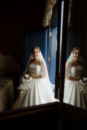 A bride in a classic white wedding gown and veil is reflected in a mirror against blue shutters in a moody setting.