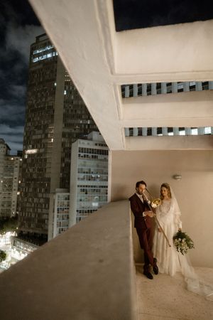 A couple in wedding attire poses in a modern architectural setting with dramatic lighting and geometric buildings in background.