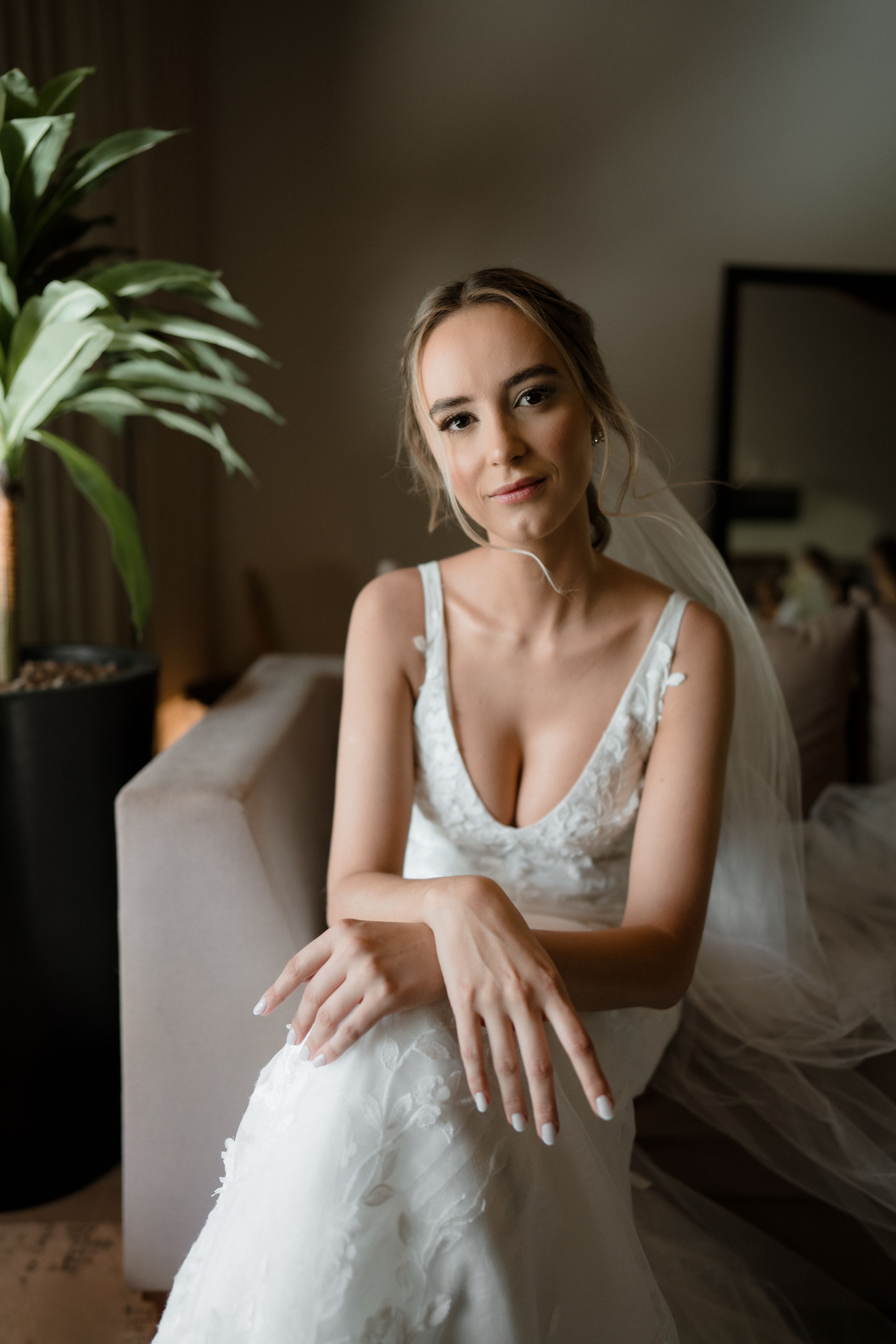 A bride in a white lace wedding dress and veil sits elegantly next to a potted plant in a dimly lit room.