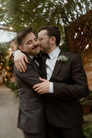 Two people in formal black suits share an intimate embrace during a wedding photoshoot in an outdoor garden setting.