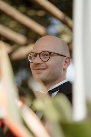 A person wearing glasses smiles while looking to the side with blurred foliage in the background.