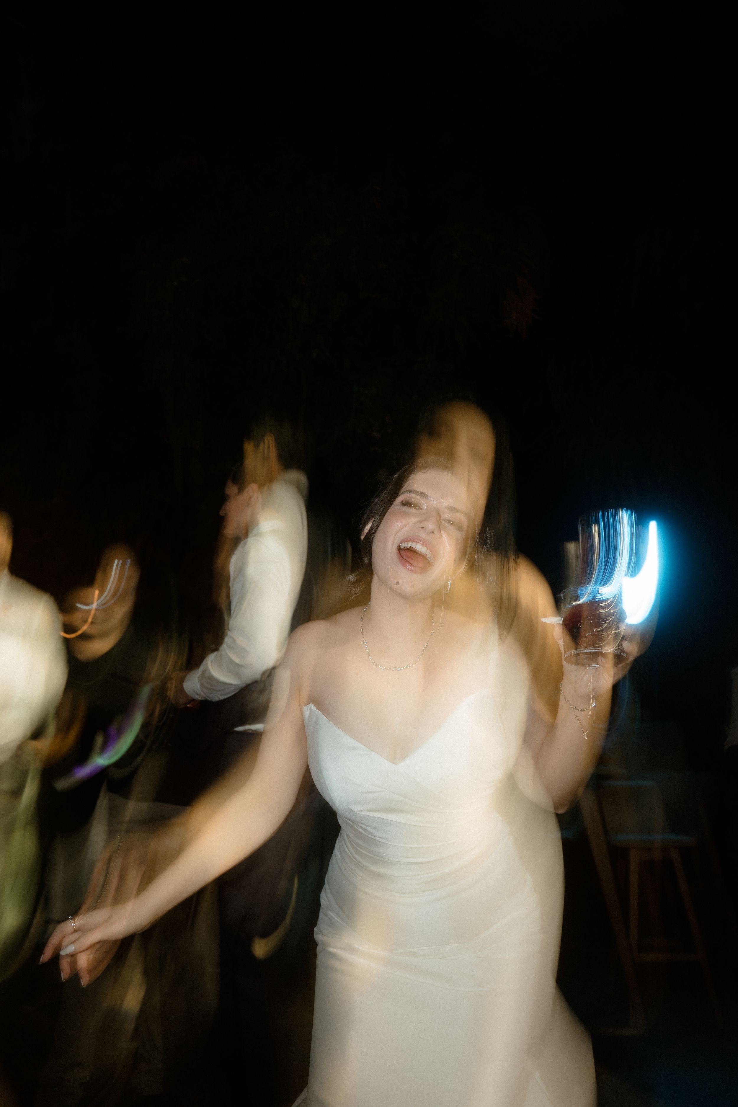 People dancing and moving energetically in a dark room with some bright lighting effects visible in the background.