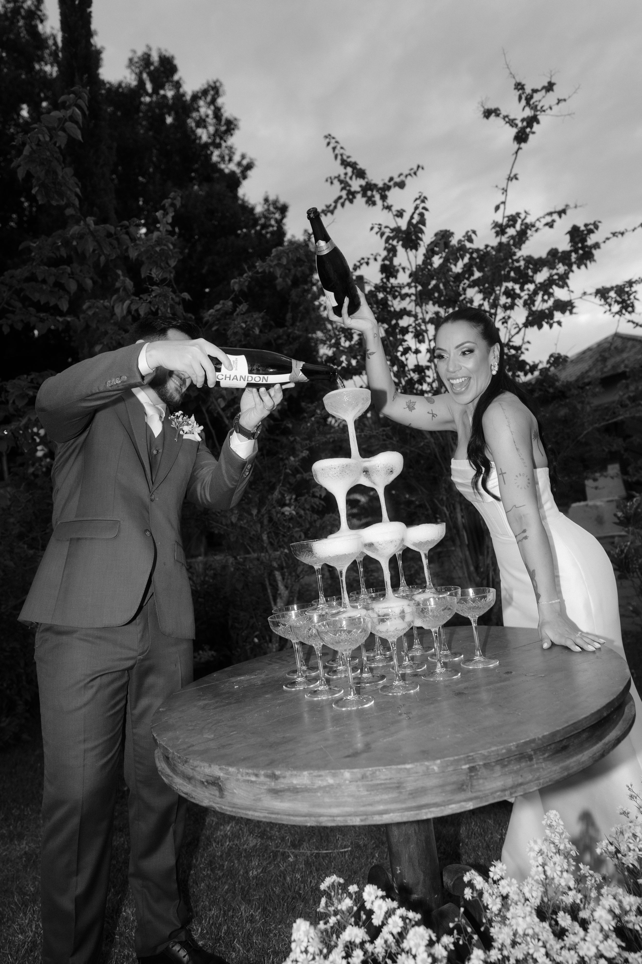 A champagne tower being carefully constructed at an outdoor wedding reception with glasses stacked in pyramid formation.