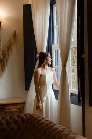 Woman in white dress stands by window with flowing curtains and macrame wall hanging in bohemian styled room.
