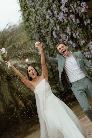 Joyful couple in wedding attire celebrating with confetti toss against floral backdrop at outdoor ceremony.