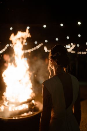 Silhouettes of people gathered around a large bonfire at night with glowing string lights in the background.