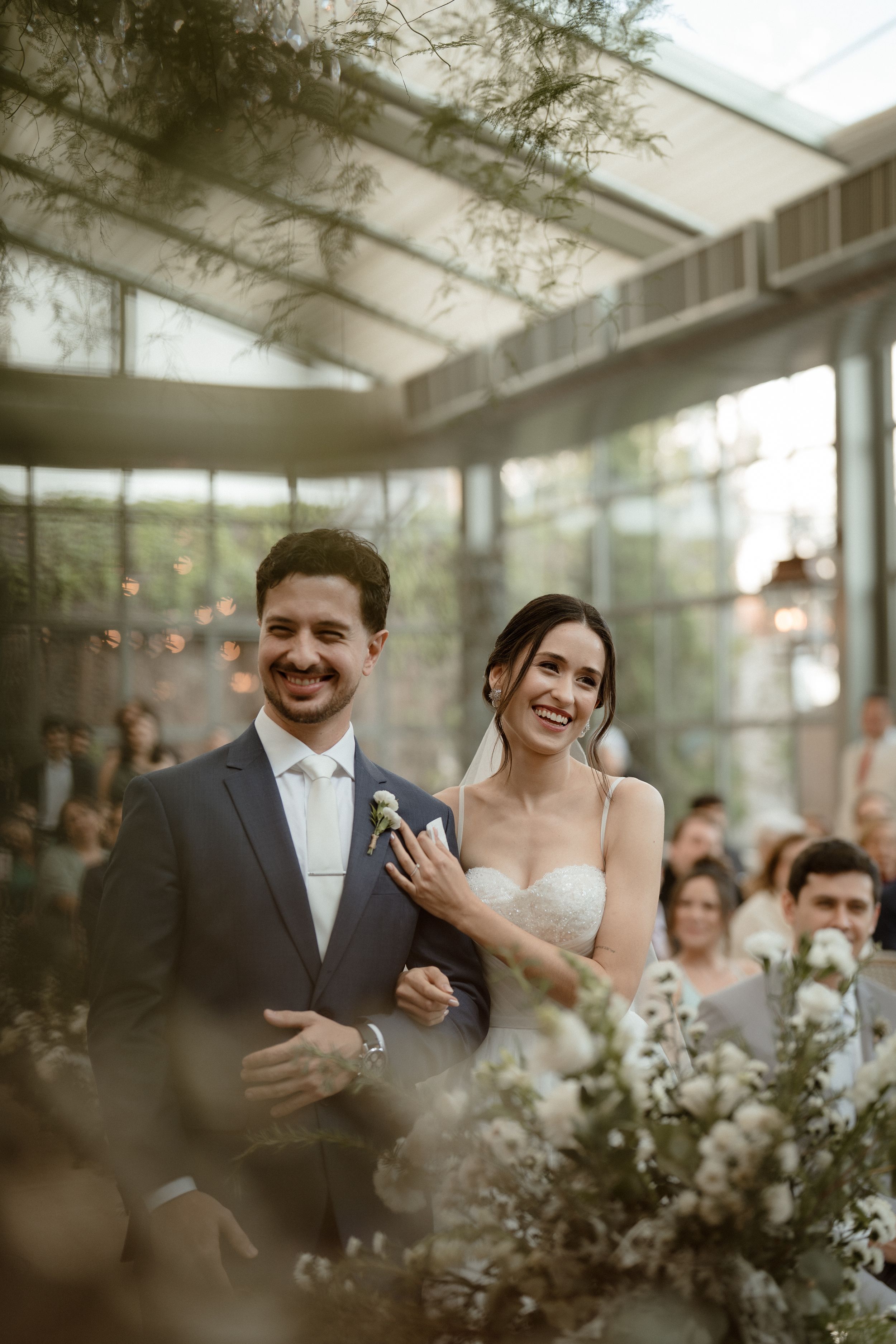 A joyful couple shares a candid moment during their greenhouse wedding ceremony with natural light streaming in.